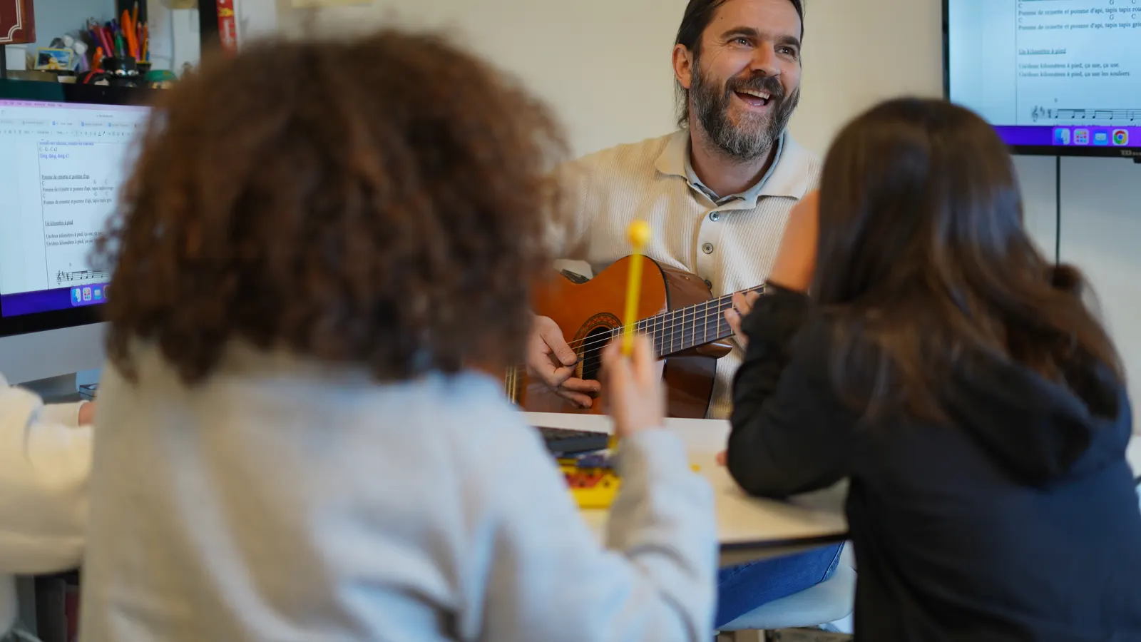 Un profesor toca la guitarra rodeado de alumnos sentados alrededor de una mesa.