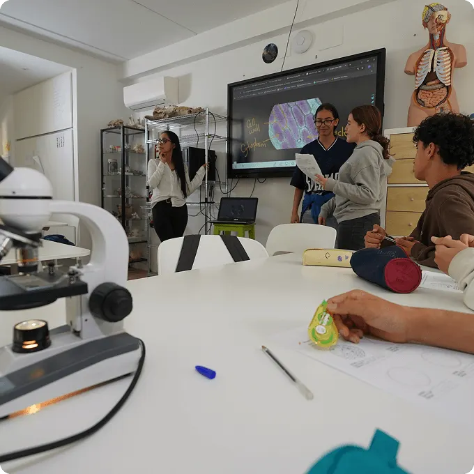Un aula con pantalla, microscopio y alumnos trabajando alrededor de una mesa ovalada.